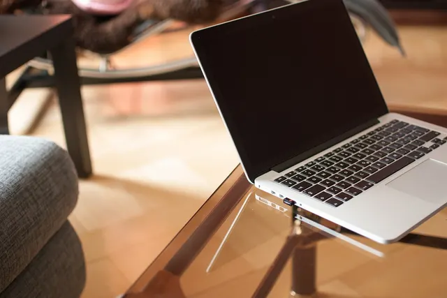 Open silver laptop with a dark screen on a glass coffee table in a sunlit living room beside a gray sofa