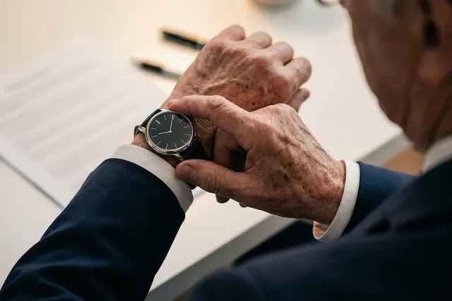 Older businessman in a navy suit checks his wristwatch at a desk with printed documents and pens