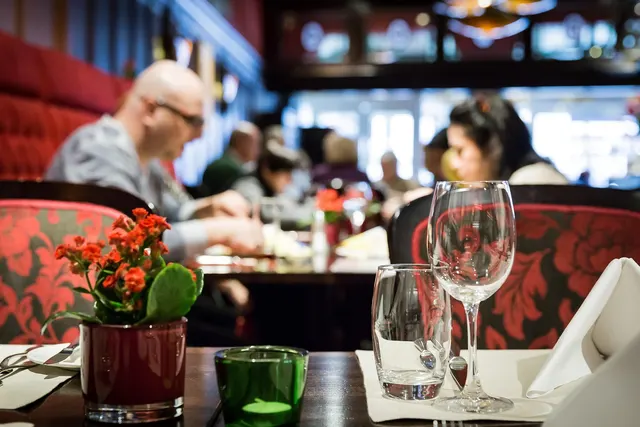 Restaurant table set with wine glass, water glass and red flowers in foreground while diners eat in the blurred background