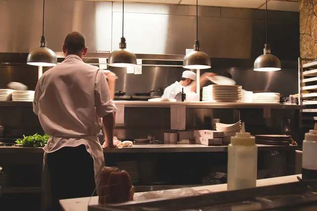 Chefs in white uniforms working in a busy stainless steel restaurant kitchen under pendant lights with stacks of plates