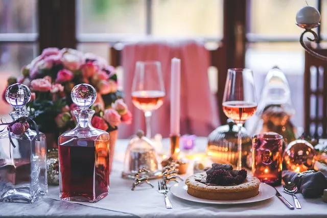 Festive dining table with rosé wine glasses, crystal decanters, pink roses, candles and a tart on a white plate