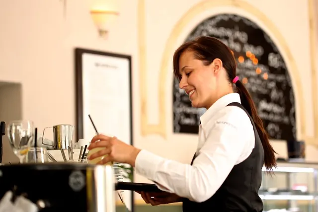 Smiling waitress in white shirt and black vest holding a tray with a drink at a cafe counter, chalkboard menu behind