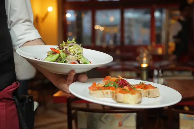 Server carrying two plated dishes through a dim dining room