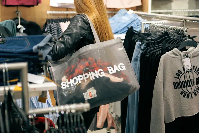 Shopper in a leather jacket carrying a clear tote of clothes past racks of hoodies and jeans in a clothing store