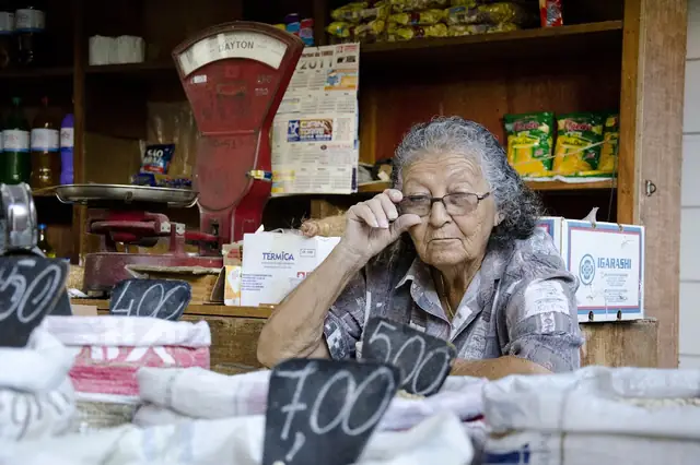 Elderly shopkeeper adjusting her glasses behind a cluttered market counter