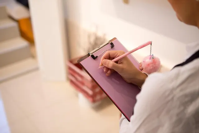 Over-the-shoulder view of a person in a white shirt writing on a pink clipboard with a pink pom-pom pen in a bright store