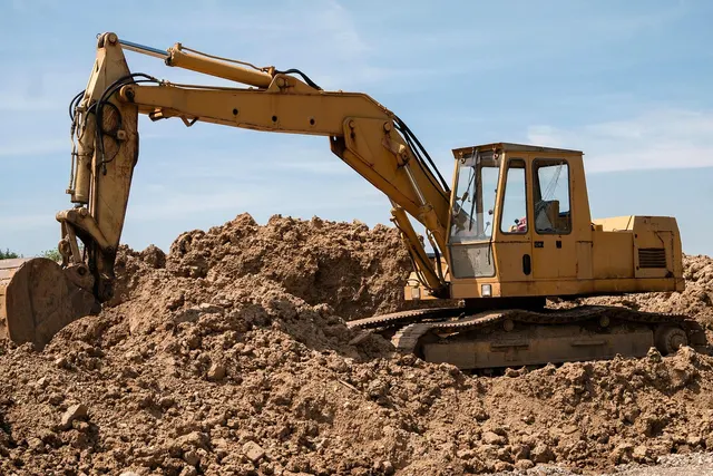 Yellow tracked excavator with its arm extended over large mounds of dirt at a construction site under a blue sky