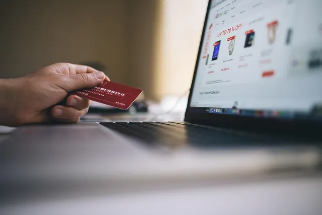 Hand holding a red payment card beside a laptop showing an online store sale page, suggesting an e-commerce checkout