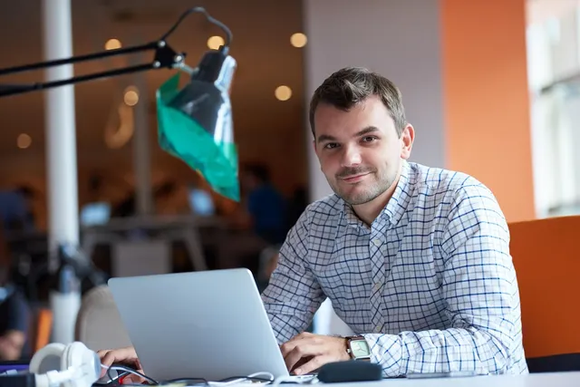 Smiling man in a checked shirt working on a laptop at a desk in a bright, busy open-plan office