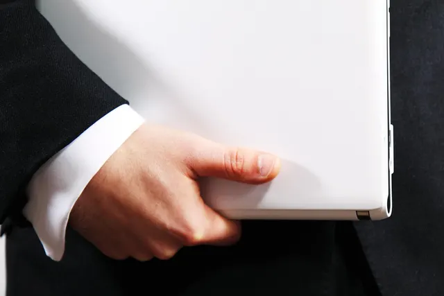 Close-up of a businessman in a dark suit and white shirt cuff carrying a closed white laptop under his arm