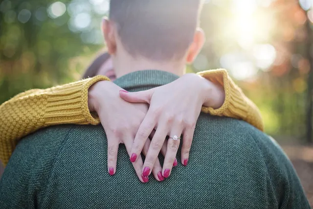 Woman with pink nails and a diamond engagement ring hugging a man in a green sweater outdoors in autumn