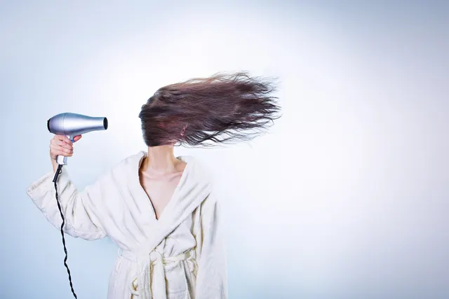 Woman in a white bathrobe holding a silver hair dryer, long brown hair blowing across her face against a pale blue wall