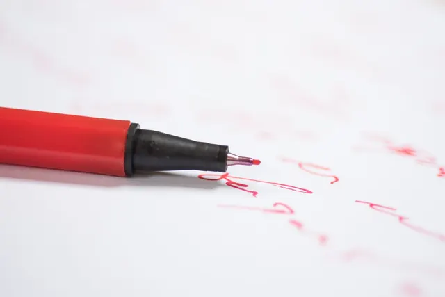 Close-up of a red fine-tip pen resting on white paper covered in blurred red handwritten marks