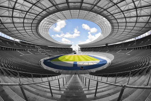 Wide-angle view of an empty sports stadium with tiered gray seats, a green pitch, a blue track and an open oval roof