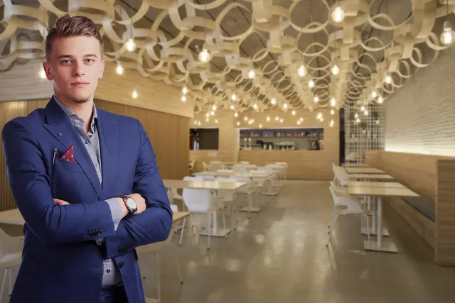 Young man in a navy suit with arms crossed standing in an empty modern restaurant with white tables and hanging bulbs