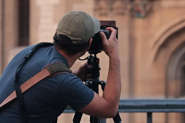 Photographer in a green cap and blue t-shirt shooting with a camera on a tripod in front of a blurred stone building