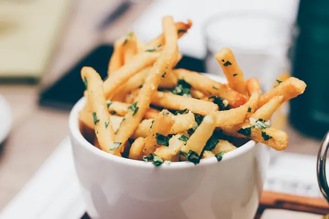White cup of golden french fries topped with chopped fresh herbs on a restaurant table, glasses blurred behind