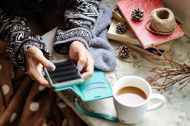 Hands holding a smartphone beside coffee and a cozy knit sweater