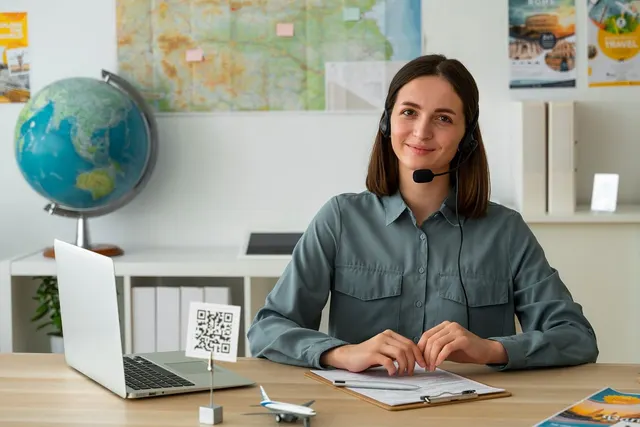 Smiling travel agent in a headset at a desk with a laptop, QR code stand and model plane, globe and map behind her