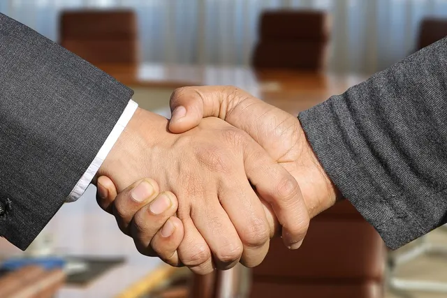 Close-up of two people shaking hands, one in a dark suit jacket, over a conference table with brown chairs