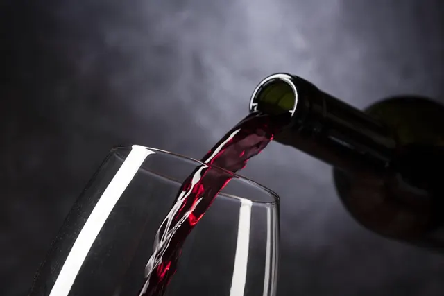 Red wine pouring from a dark green bottle into a clear wine glass against a smoky gray background