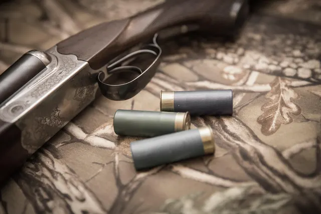 Three shotgun shells beside an engraved side-by-side shotgun resting on camouflage fabric