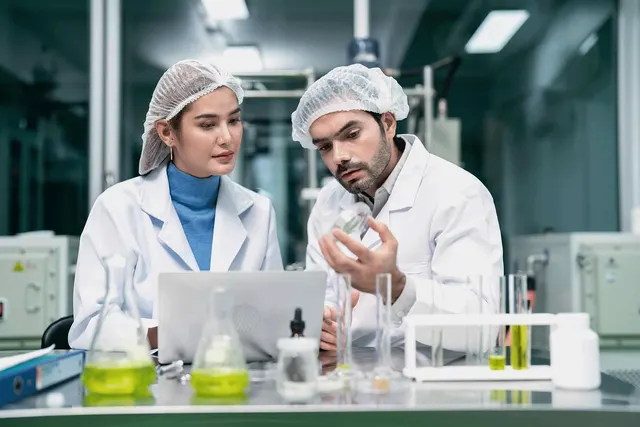 Two lab workers in hairnets and white coats examine a vial beside a laptop, flasks and test tubes of yellow liquid