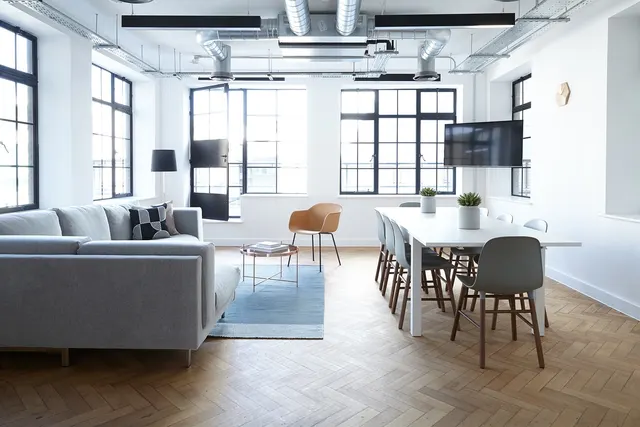 Bright loft office with grey sectional sofa, white meeting table and chairs, wall-mounted TV and tall industrial windows