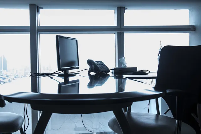 Empty office desk with a computer monitor and desk phone in front of large windows overlooking a city skyline