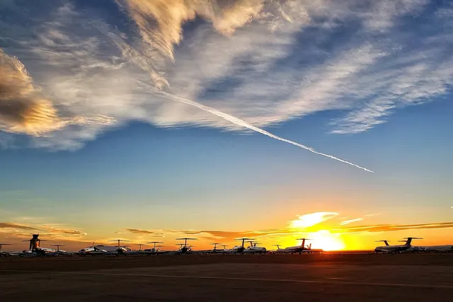 Rows of parked private jets on an airfield at sunset under a blue sky with wispy clouds and a contrail