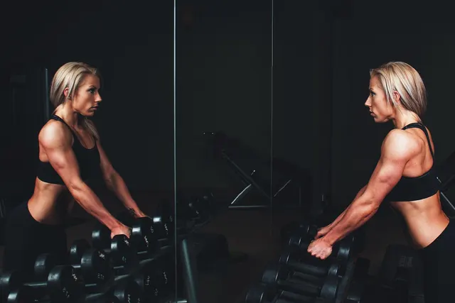 Muscular blonde woman in black sportswear gripping a dumbbell rack, facing her reflection in a dark gym mirror