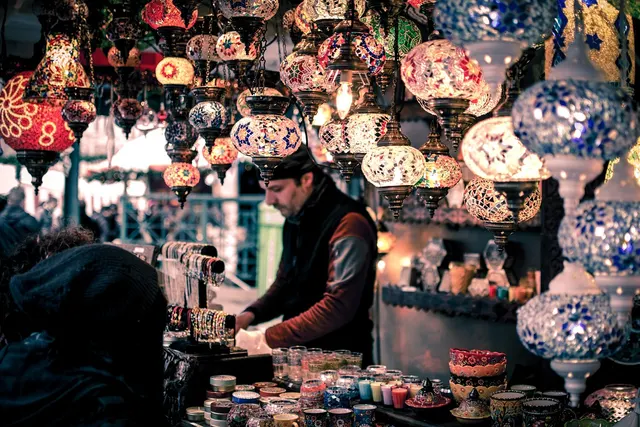 Vendor at a market stall lit by hanging mosaic lamps, with bracelets, candle holders and bowls displayed for sale