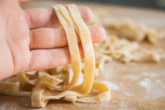 Hand lifting fresh flour-dusted homemade noodles above a pile of cut pasta ribbons on a wooden board