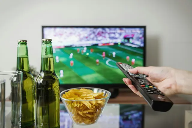Hand pointing a TV remote at a live soccer broadcast, with two green beer bottles and a bowl of chips in the foreground