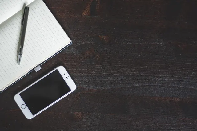 White smartphone with a blank screen beside an open lined notebook and silver pen on a dark wooden desk