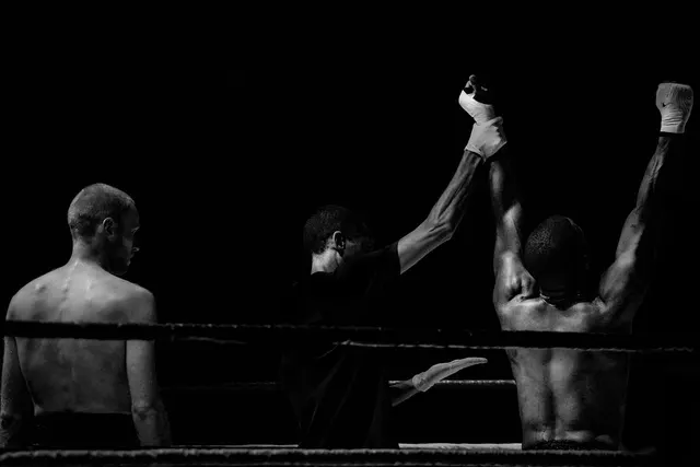 Black-and-white photo of a referee raising a boxer's taped fist in victory as the defeated fighter looks away in the ring