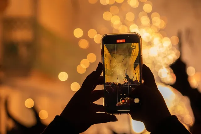 Silhouetted hands recording a night festival fireworks display on a smartphone, with glowing golden bokeh lights behind