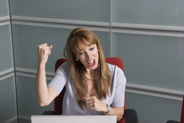 Excited businesswoman pumping her fist in celebration while looking at a laptop in a glass-walled office