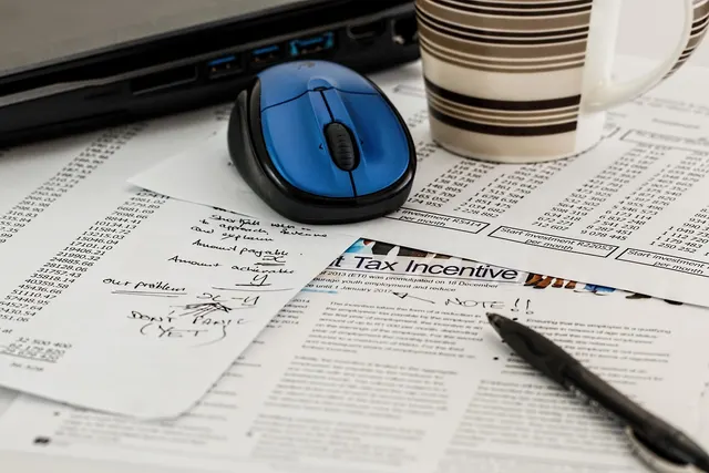Printed financial spreadsheets and a tax document with handwritten notes beside a laptop, blue mouse, pen and coffee mug