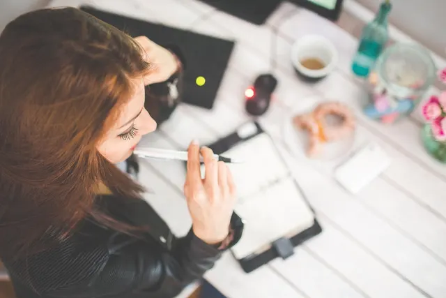 Overhead view of a woman with a pen reviewing an open planner at a white desk with coffee, a mouse and a donut