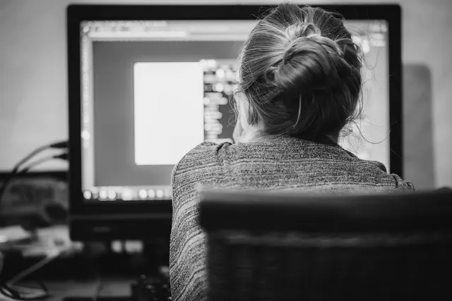 Black and white photo of a woman with her hair in a bun, seen from behind, working at a desktop computer monitor