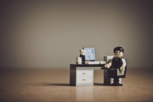 Toy minifigure with a worried expression sitting at a miniature office desk with a computer monitor and lamp