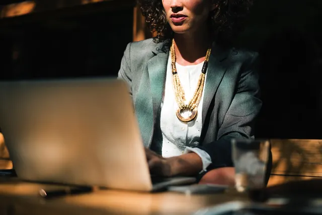 Businesswoman in a gray blazer and beaded necklace typing on a laptop at a wooden table in warm sunlight