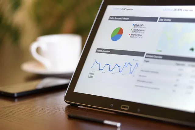 Tablet on a wooden desk showing an analytics dashboard with pie and line charts, beside a coffee cup, phone and stylus
