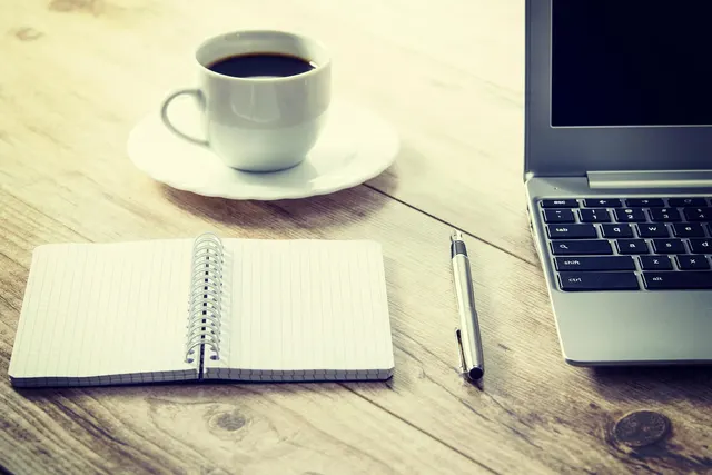 Laptop on a wooden desk beside an open spiral grid notebook, silver pen and a cup of black coffee on a saucer