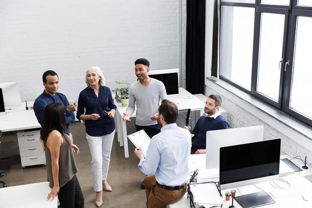 Six colleagues talk in a bright office with desktop monitors, one man holding a clipboard