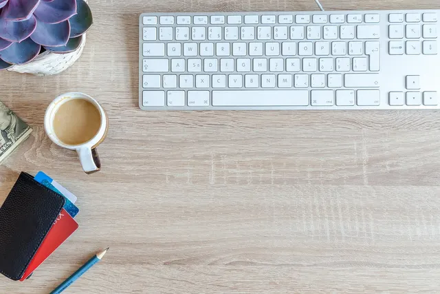 Overhead view of a wooden desk with a white keyboard, coffee cup, succulent, wallet with payment cards, cash and a pencil