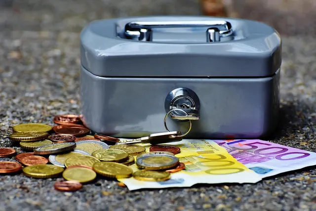 Gray metal cash box with key in the lock beside scattered euro coins and 200 and 500 euro banknotes on pavement