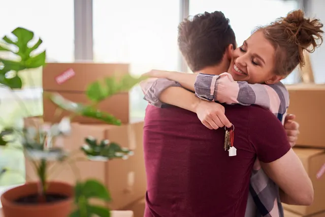 Smiling couple hugging among moving boxes, woman holding house keys with a house-shaped keychain