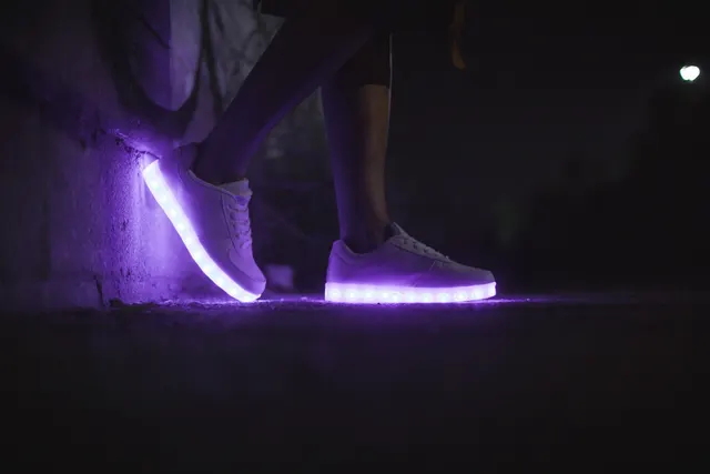 Person wearing white LED light-up sneakers glowing purple at night, one foot propped against a wall